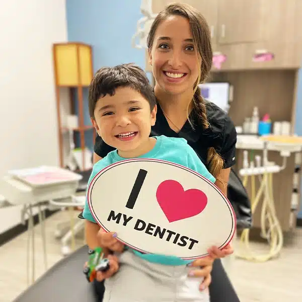 Happy young girl patient at Edna dentistry. The girl is taking a photo with the doctor holding in hand a card which is written: "I love my dentist".
